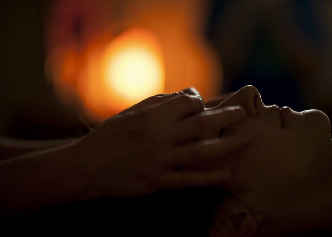 Mains posées avec douceur sur le visage d'une personne allongée, à la lueur d'une bougie, pendant une séance de magnétisme